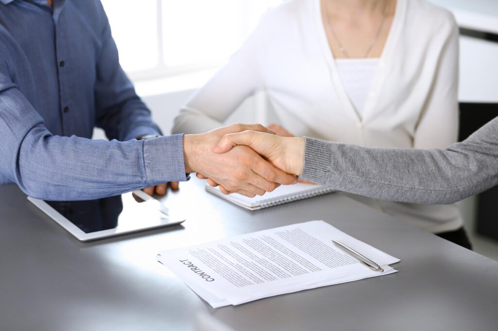 Two individuals shaking hands over a desk, representing a partnership at All West Law Group.