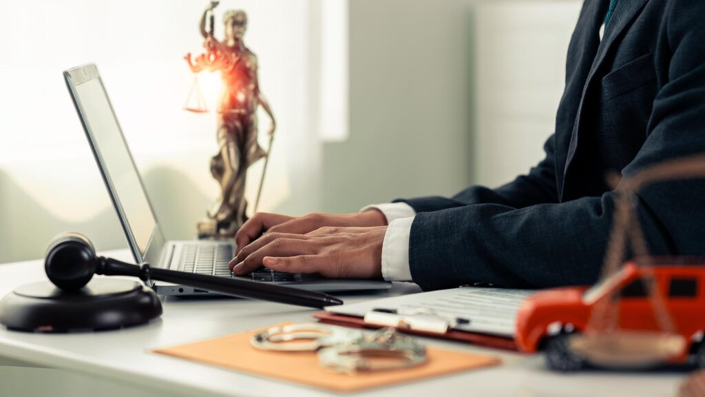 A person in a suit types on a laptop beside a desk with a Lady Justice statue, scales glowing. Nearby are a gavel, clipboard, handcuffs, and a toy car.