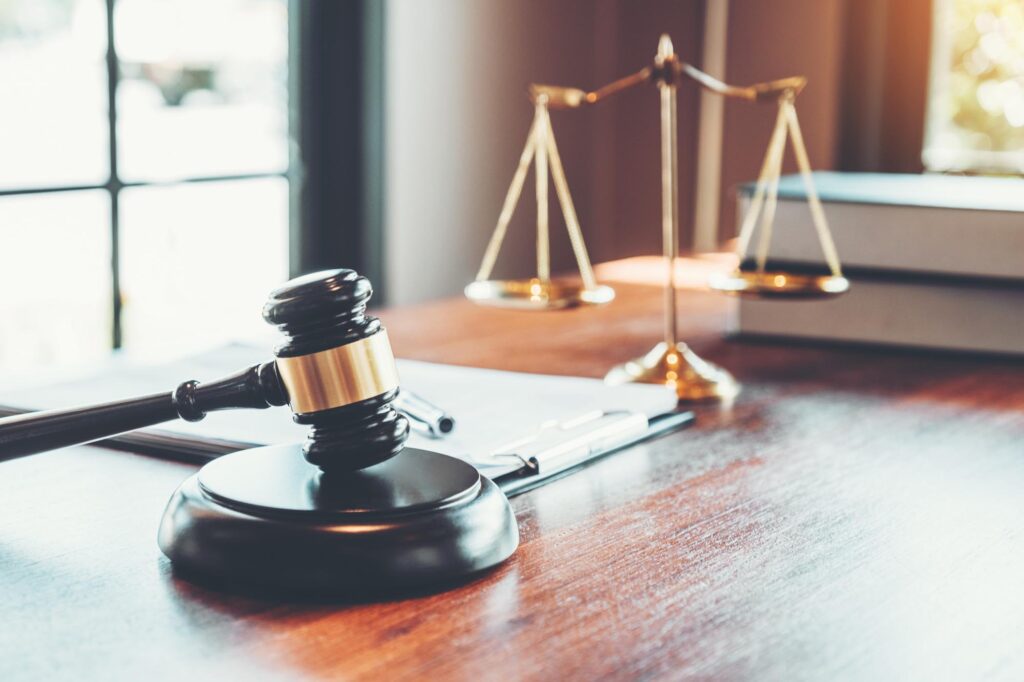 A wooden judge's gavel rests on a desk beside a balanced scale, symbolizing justice. Books and papers are in the background, suggesting a legal setting.