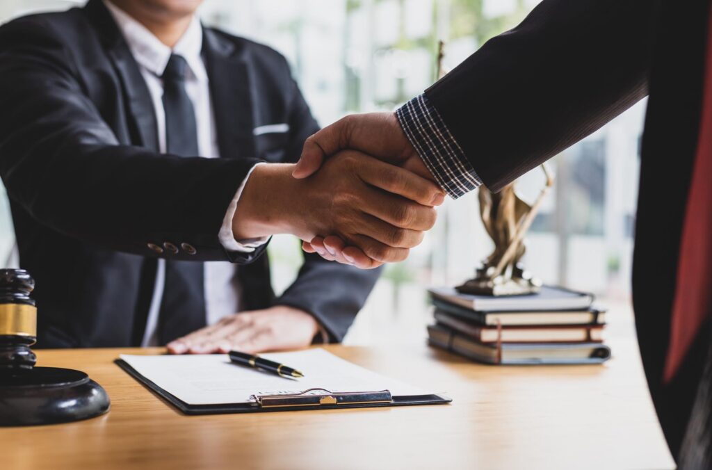 Two people in business attire shake hands over a table with a contract, pen, gavel, and statue, symbolizing agreement or partnership in a legal setting.