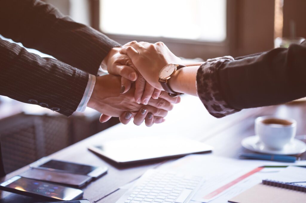 Close-up of four hands in business attire stacked together over a desk, symbolizing teamwork and collaboration. Papers and phones are in the background.