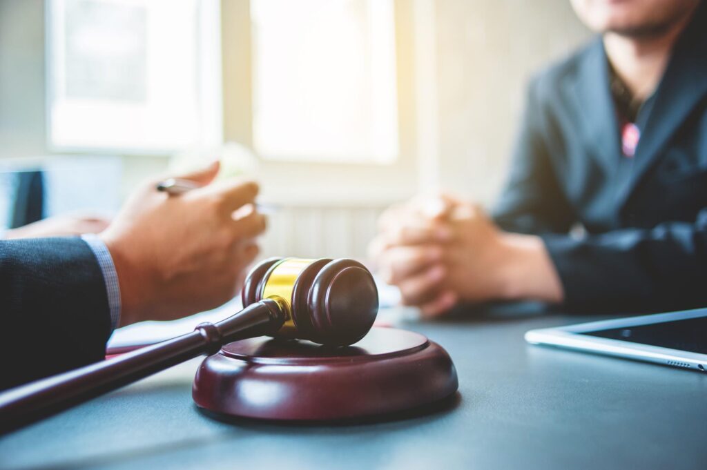 Close-up of a judge's gavel on a desk with two people in suits having a discussion. The setting suggests a formal, legal tone.
