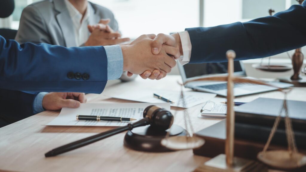 Handshake between two people in business attire over a table with legal documents, a gavel, and scales of justice, conveying agreement and professionalism.