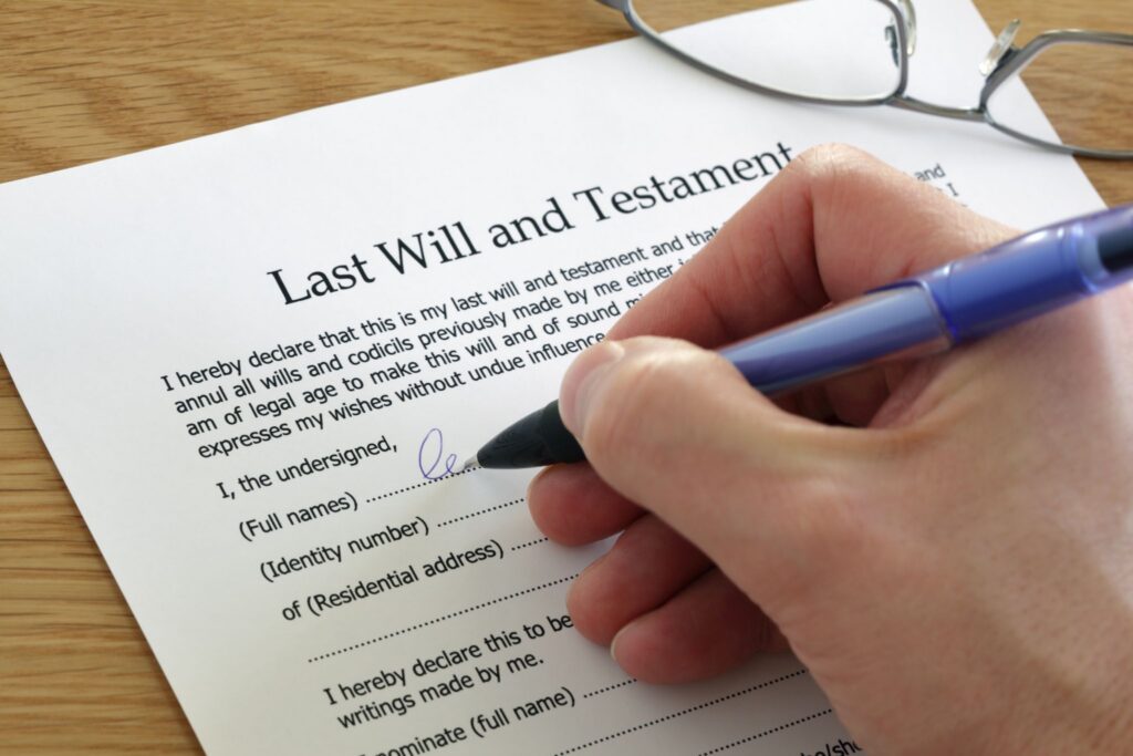 A close-up of a person's hand using a blue pen to fill out a Last Will and Testament document on a wooden desk.