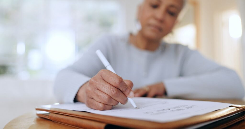 A close-up of a woman's hand signing a document with a white pen on a wooden table.