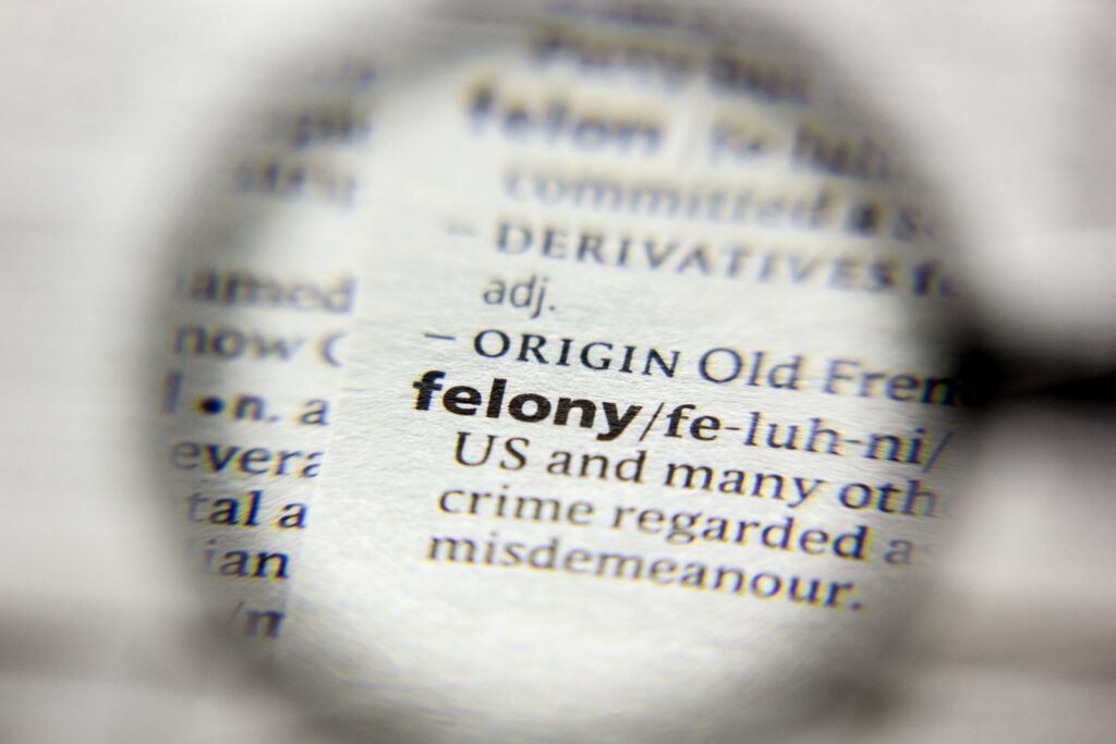 A magnifying glass focusing on the word felony and its definition in a dictionary.