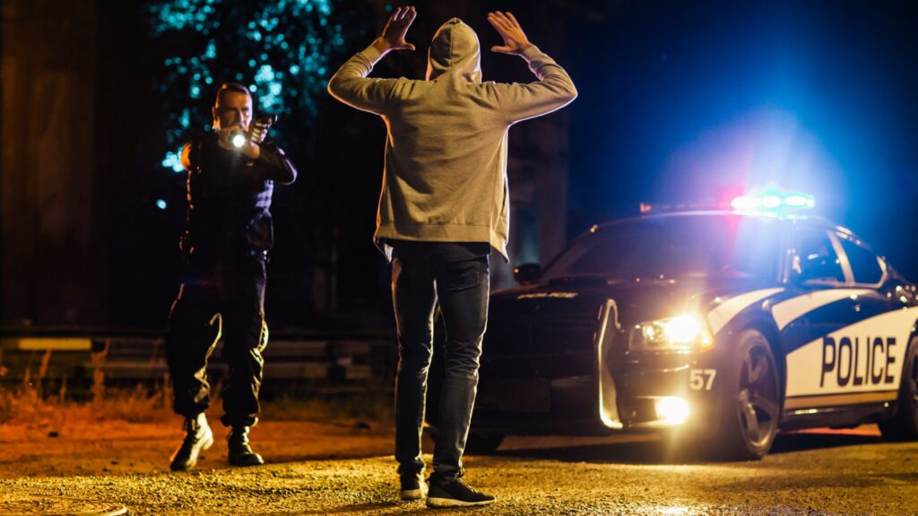 A police officer pointing a weapon at a suspect with their hands raised at night in front of a police car with flashing lights.