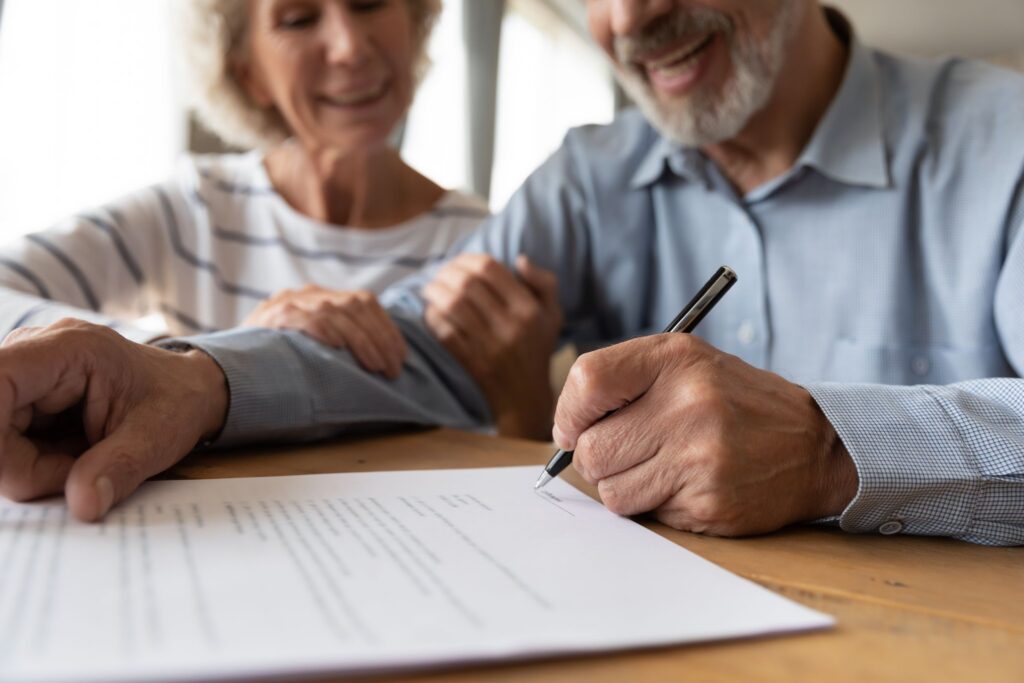 A senior couple smiling while the man signs a document at a table.