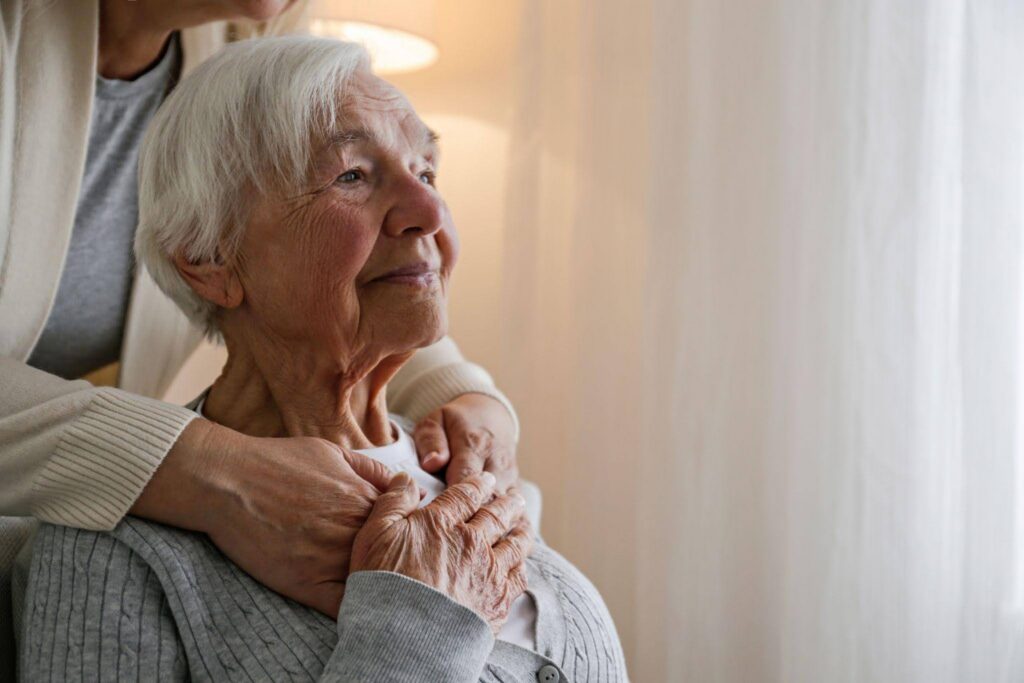 An elderly woman smiles softly by a bright window as a caregiver embraces her from behind