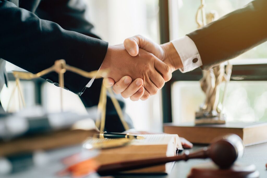 Two individuals in suits shaking hands over a law desk with a brass scale, gavel, and books in the foreground.