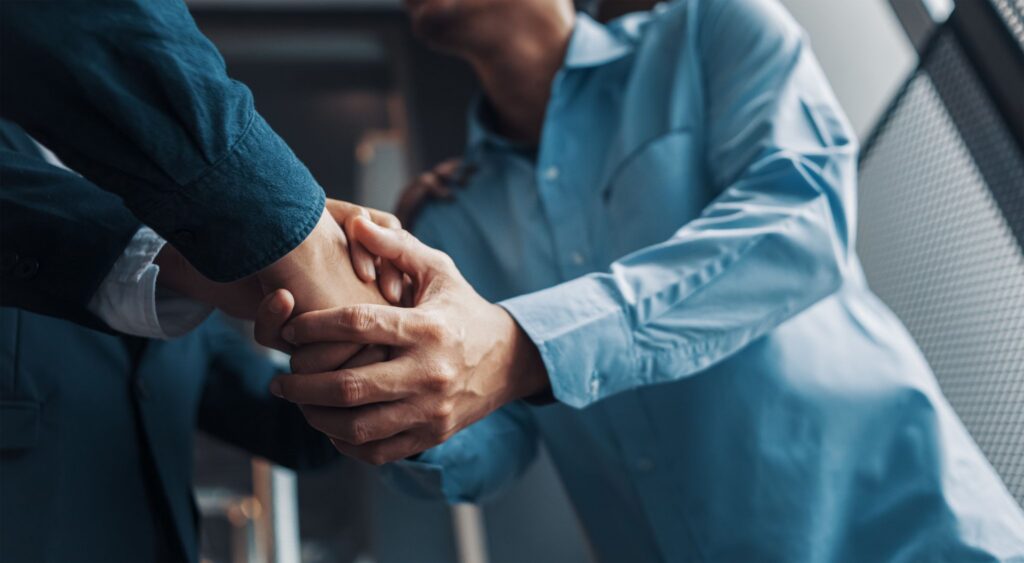 Two professional men in button-down shirts offering a firm, supportive two-handed handshake.