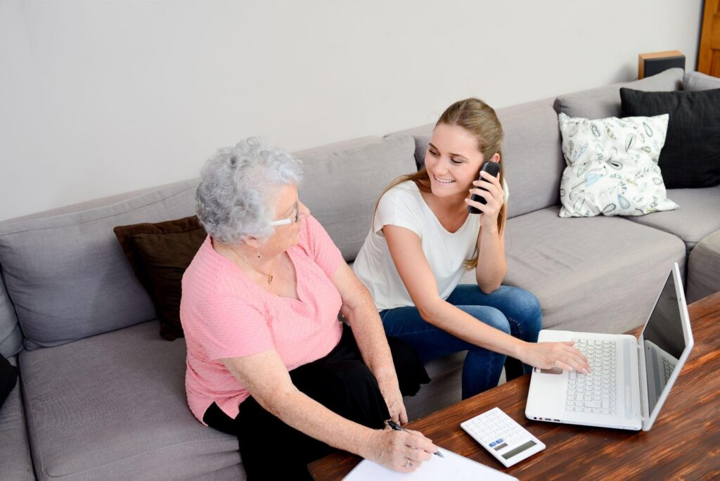 Smiling caregiver helps an older woman use a laptop and smartphone